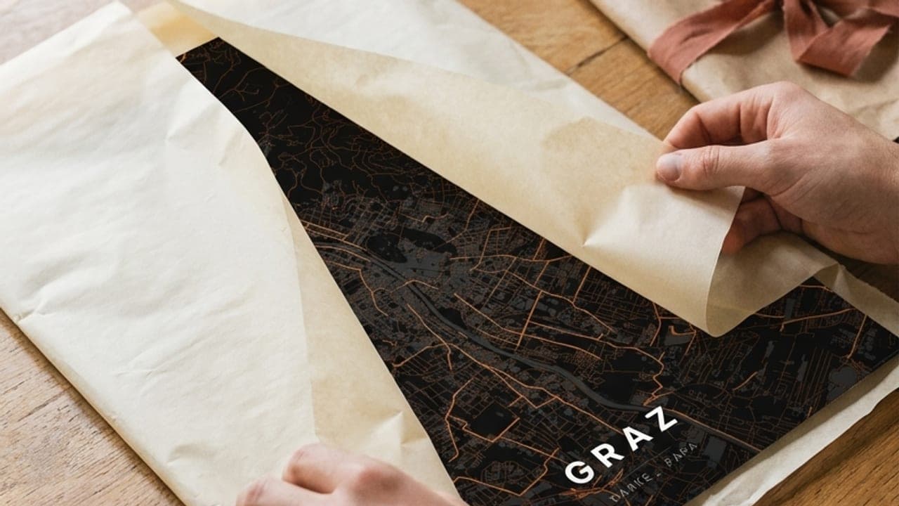 Hände packen ein dunkles Graz-Kartenposter aus Geschenkpapier aus